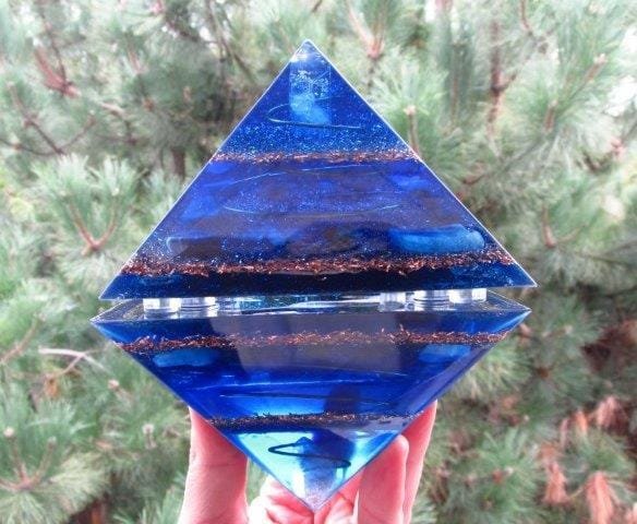 Two blue orgone pyramids held in an octahedron shape in front of a tree.