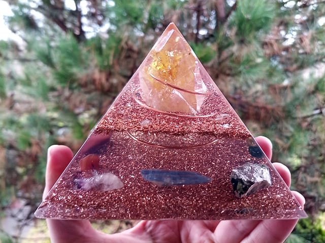 Abundance orgone pyramid in front of a tree with blue kyanite and emerald crystals.