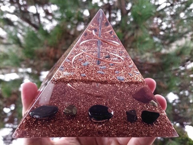 Blue kyanite orgone pyramid with shungite protection crystal, held in front of tree.