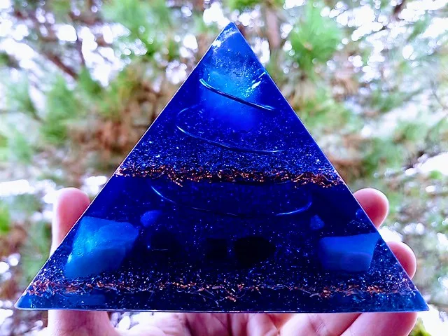 Blue orgonite pyramid with blue crystals held in front of a tree.