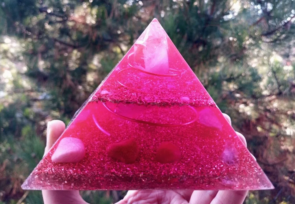 Divine love orgone pyramid, which is pink, being held in front of a pine tree.