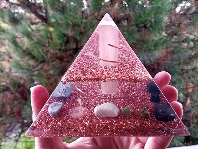 Selenite orgone pyramid with chakra crystals, being held in front of a tree.